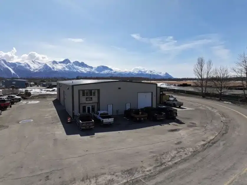 A large warehouse with a few trucks parked in front is surrounded by a wide paved area. In the background, snow-capped mountains rise under a clear blue sky. Trees line the road adjacent to the building.