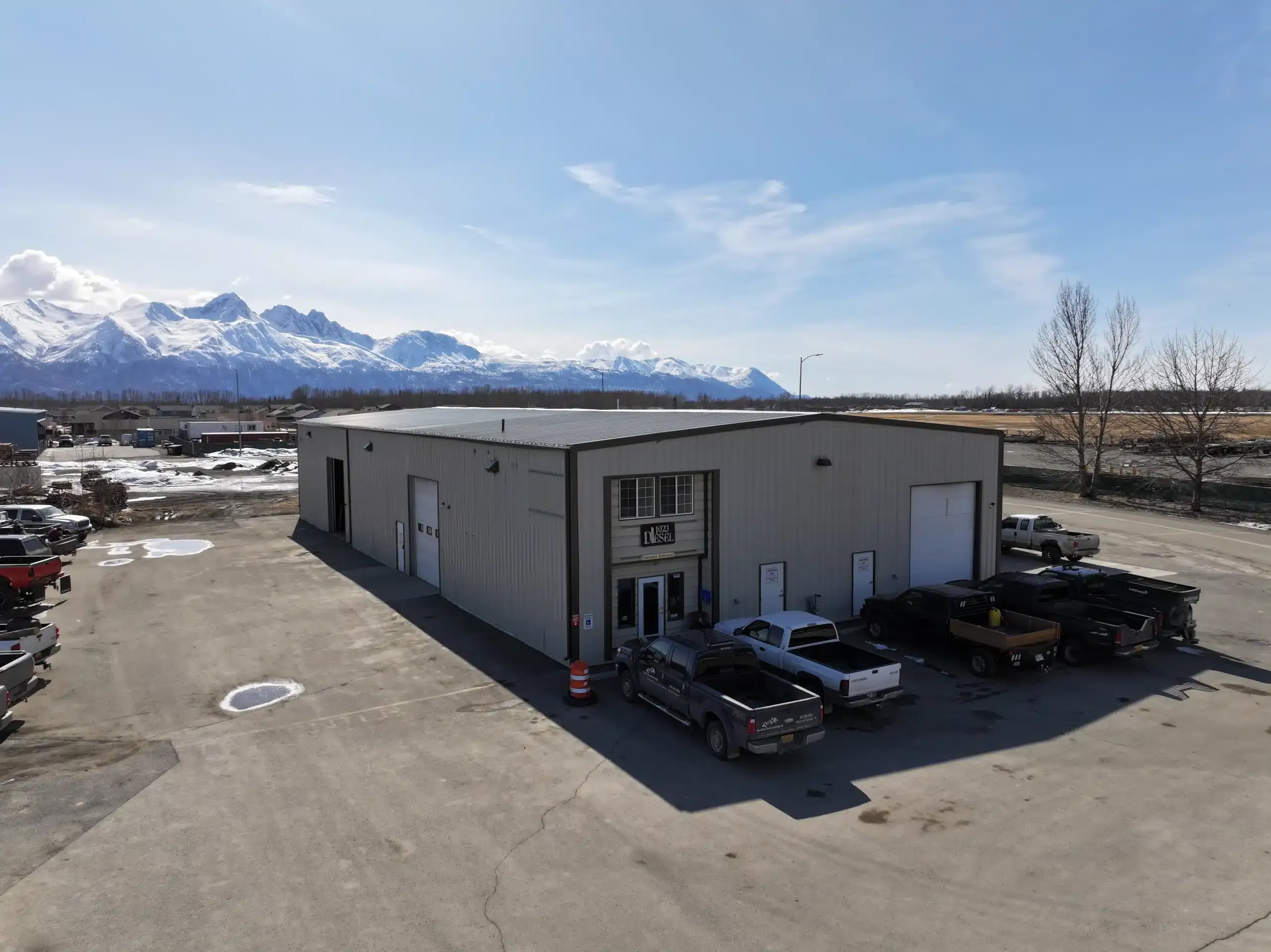 A large industrial building with a gray exterior, surrounded by parked trucks on a spacious lot. Snow-capped mountains and a clear blue sky are visible in the background.