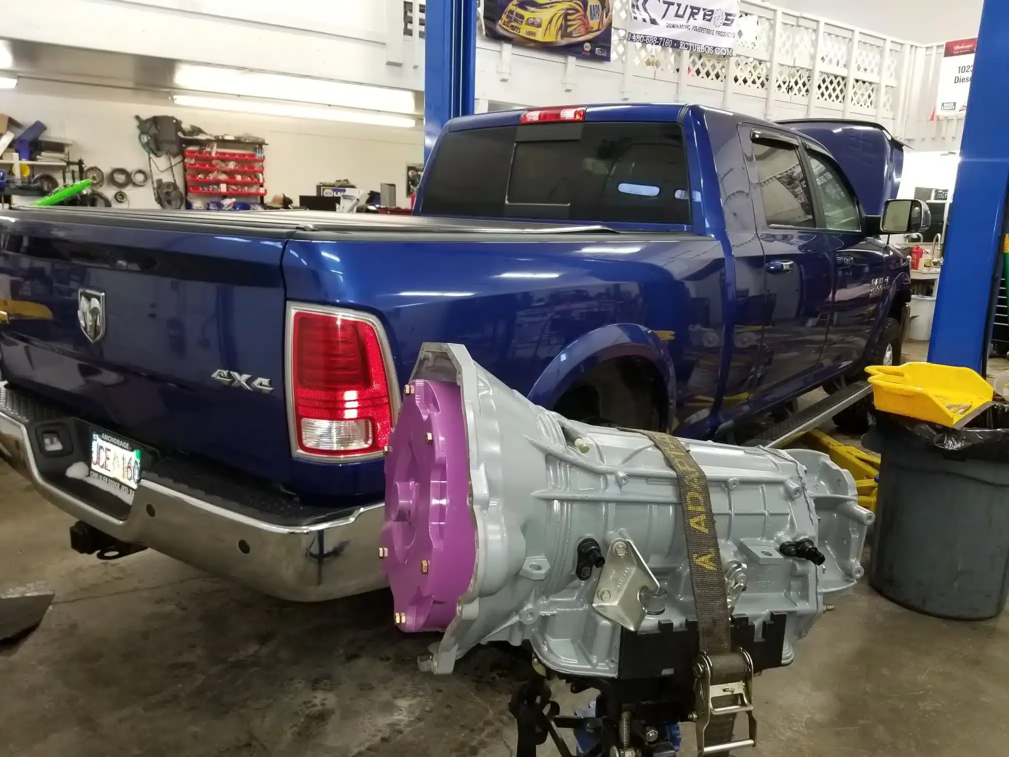 A blue pickup truck is parked inside a garage. In the foreground, there is a large transmission unit on a stand. The garage has tools and equipment visible in the background.