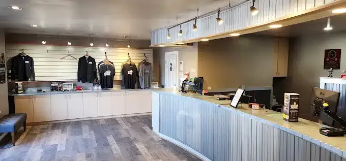 A modern retail store interior with a curved counter, computer, hanging lights, and merchandise on shelves and hangers. The floor is wooden, and the walls have a mix of light and neutral tones. Various clothing items are displayed on the left.