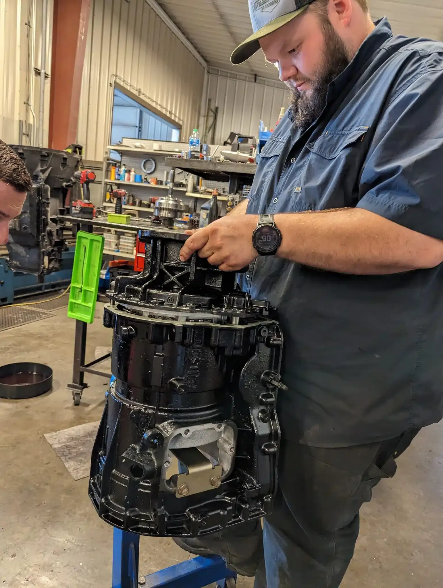 A man wearing a cap and black shirt is working on a large black mechanical component in a workshop. Shelves with tools and parts are visible in the background under fluorescent lighting. Another person is partially visible on the left.