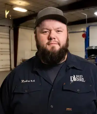 A bearded technician in a 1023 Diesel cap and work shirt standing in the shop.
