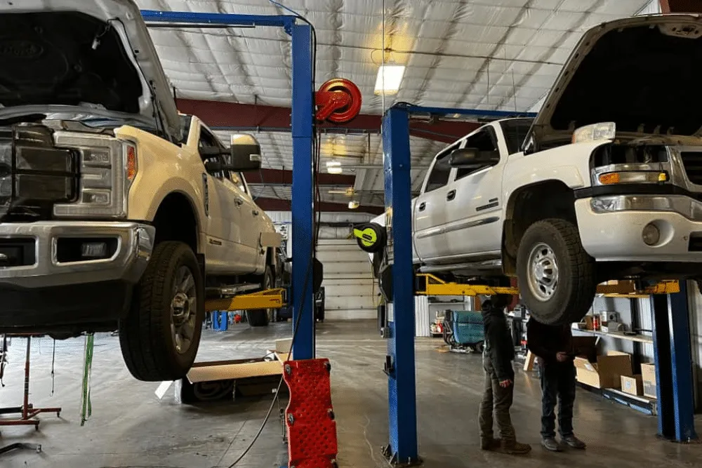 Two heavy-duty diesel trucks lifted in a spacious repair shop at 1023 Diesel & Fleet in Palmer, AK, with mechanics inspecting the vehicles for maintenance and repairs.