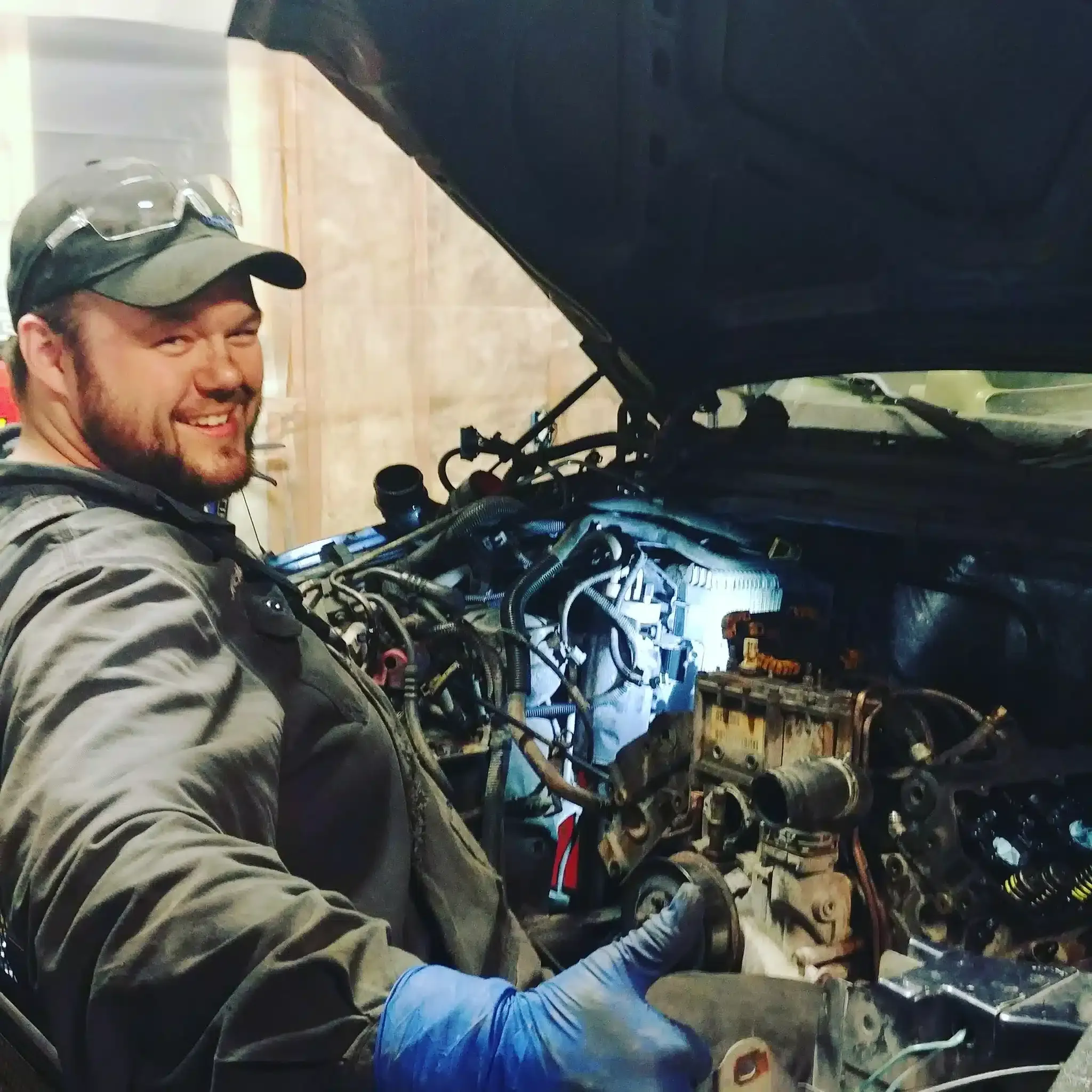 A man in a cap and safety glasses is smiling while working on a car engine. The hood is open, and he is giving a thumbs-up with his gloved hand, indicating satisfactory progress or completion of the task.