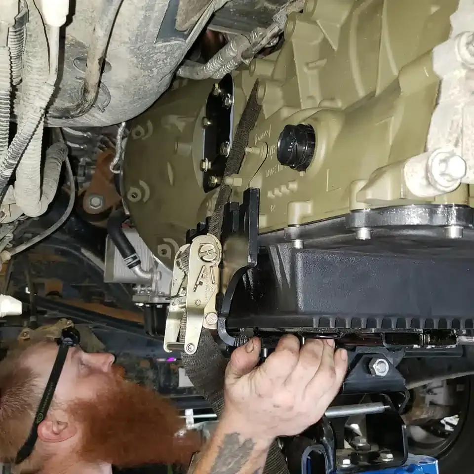 A person with a beard is working under a vehicle, focusing on a component, possibly the transmission, with a strap and tool for support. The person is wearing a headlamp while lying beneath the vehicle. The area is cluttered with cables and tubes.