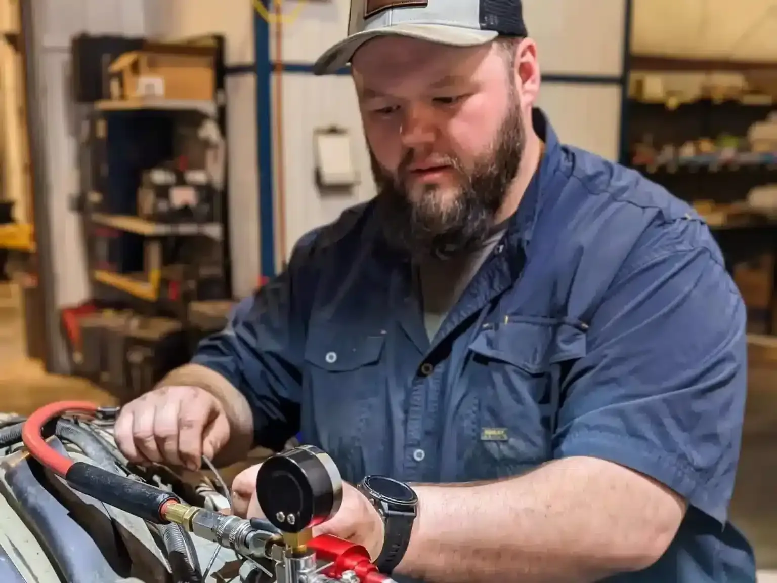 A man with a beard, wearing a cap and a blue shirt, is focused on repairing equipment in a workshop. He is adjusting parts of the machinery with various tools around him. The background shows shelves with tools and equipment.