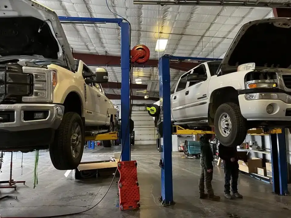 Two white pickup trucks are elevated on hydraulic lifts in a spacious garage. A person in work attire stands under one of the trucks, while another stands nearby. The garage is well-lit with visible equipment and tools in the background.