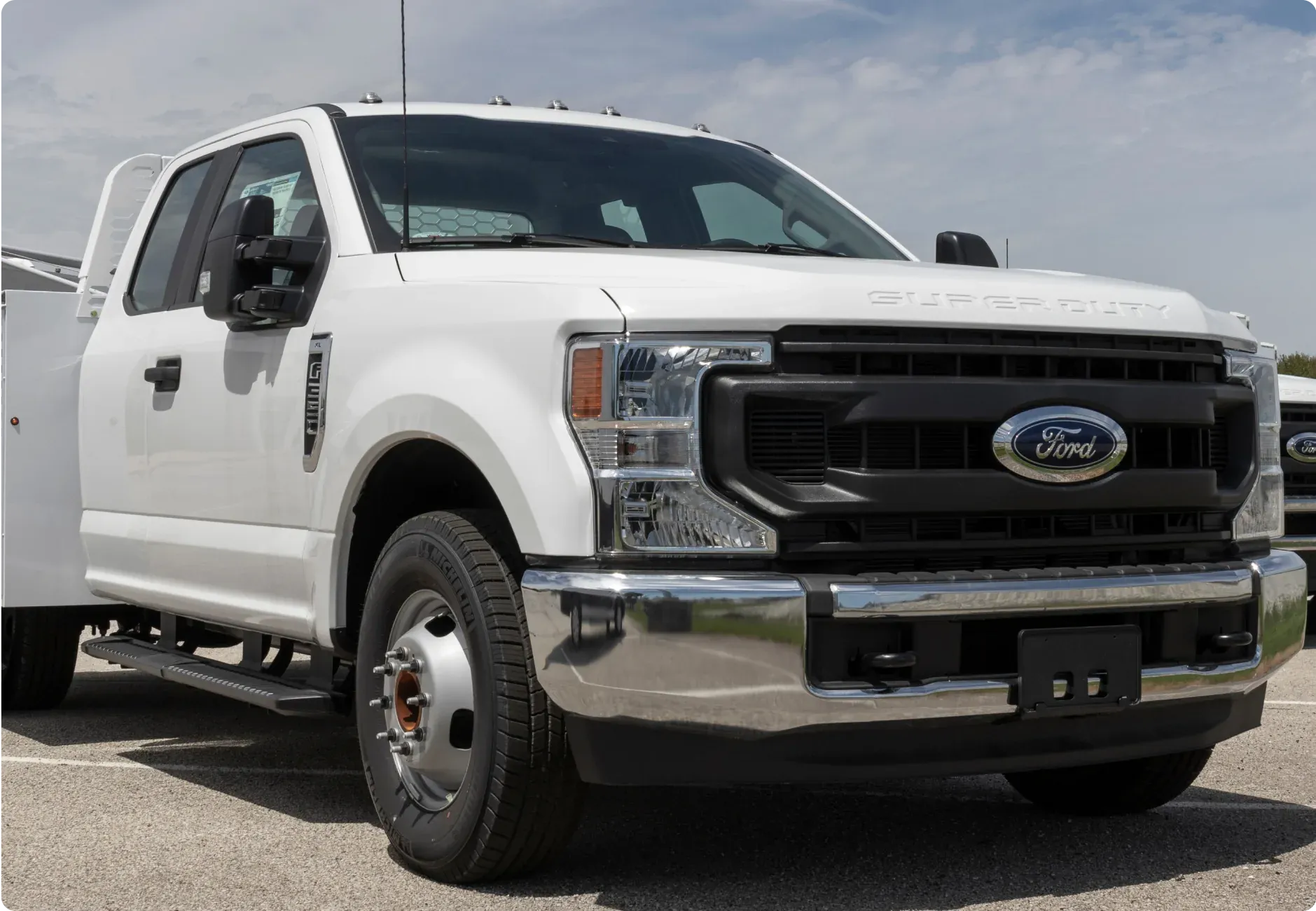 1023 Diesel & Fleet: a white Ford Super Duty truck parked outdoors on a sunny day, seen from a low angle with its front grille and headlights in view.