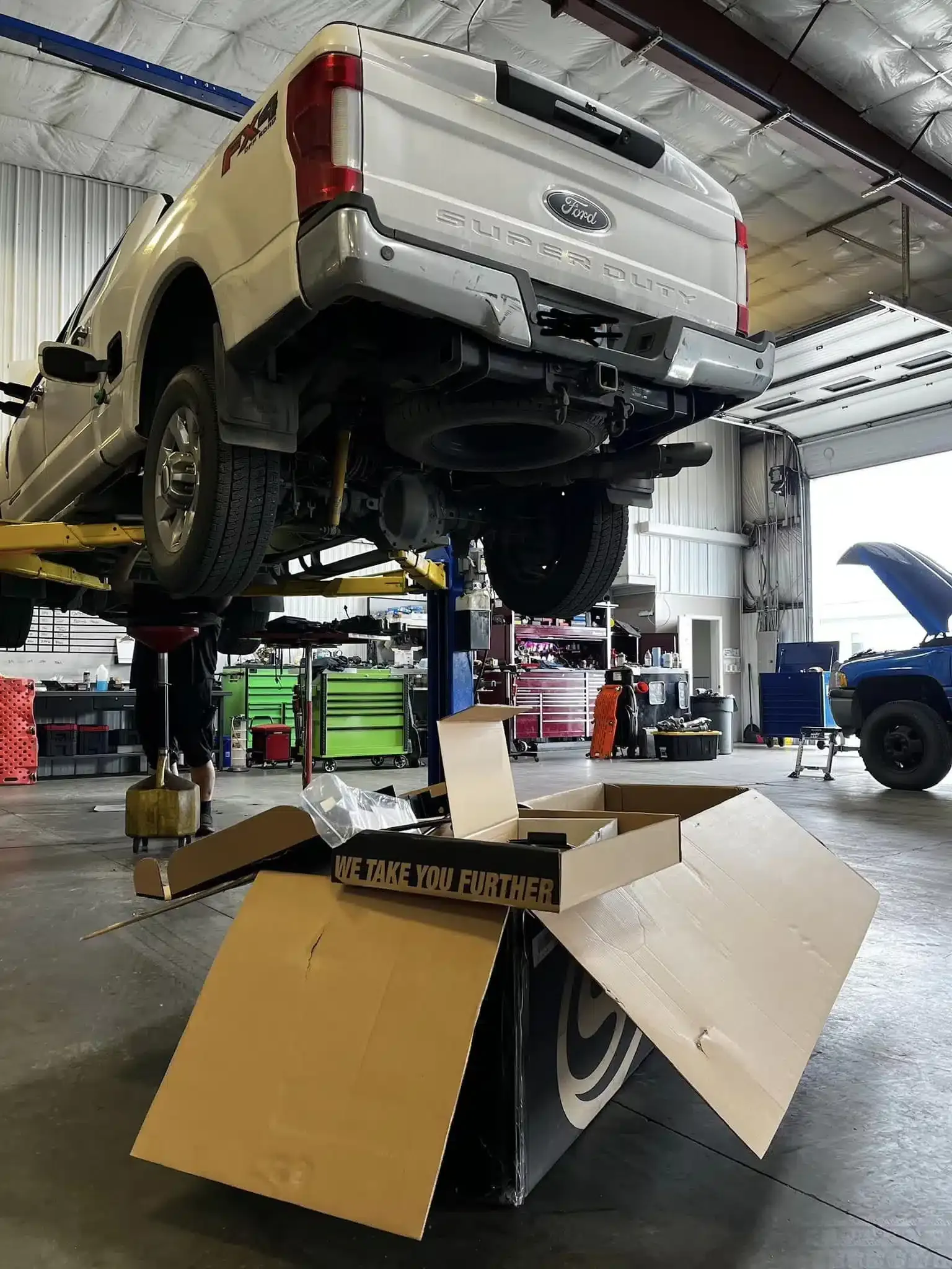 A white pickup truck on a hydraulic lift in an auto repair shop. Various tools and equipment are visible, with a large cardboard box labeled We Take You Further in the foreground. Another vehicle is being serviced in the background.
